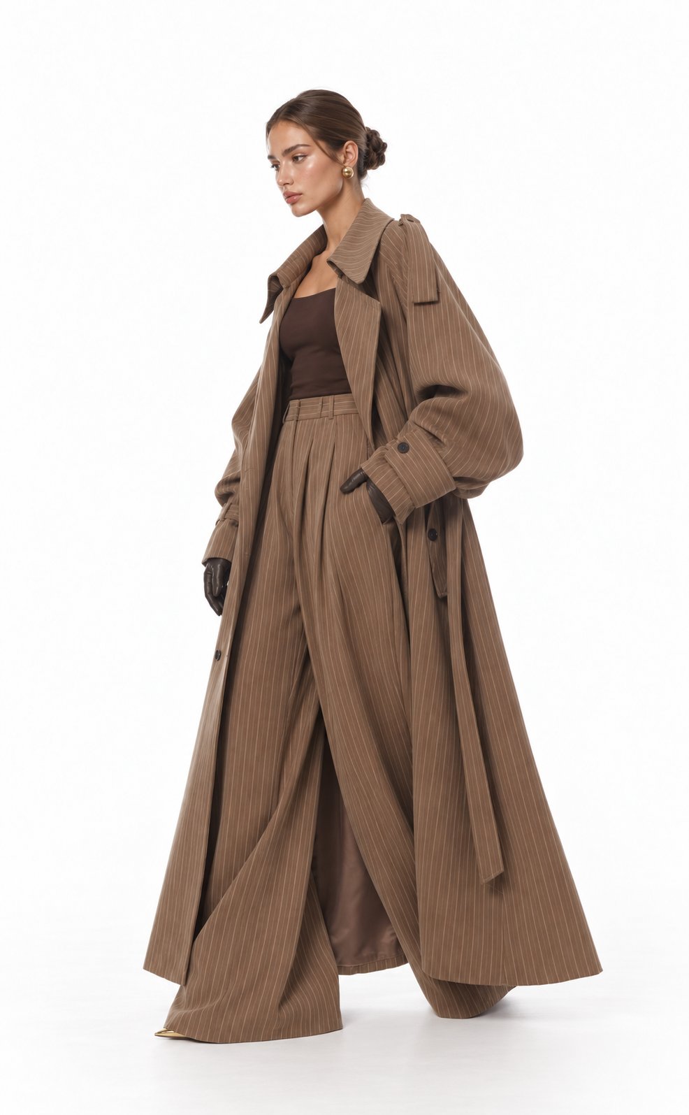 A model is shown from the side wearing a long, wide-leg brown pleated coat over a matching brown top and trousers, set against a plain white studio background.