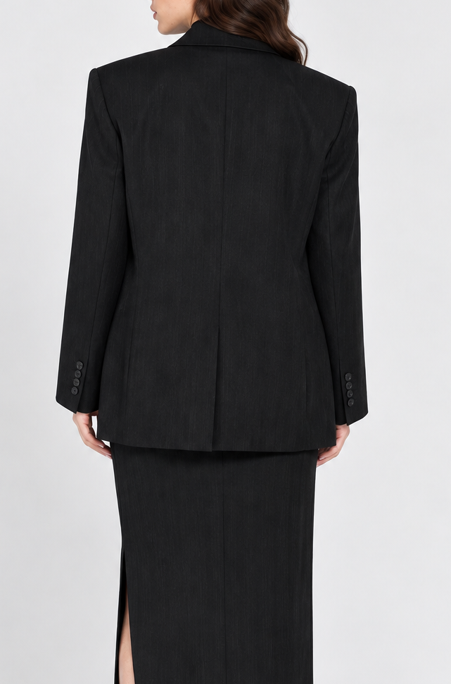 A woman shown from the back wearing a black tailored blazer and a long black skirt with a front slit, paired with black heels, against a light gray studio background.