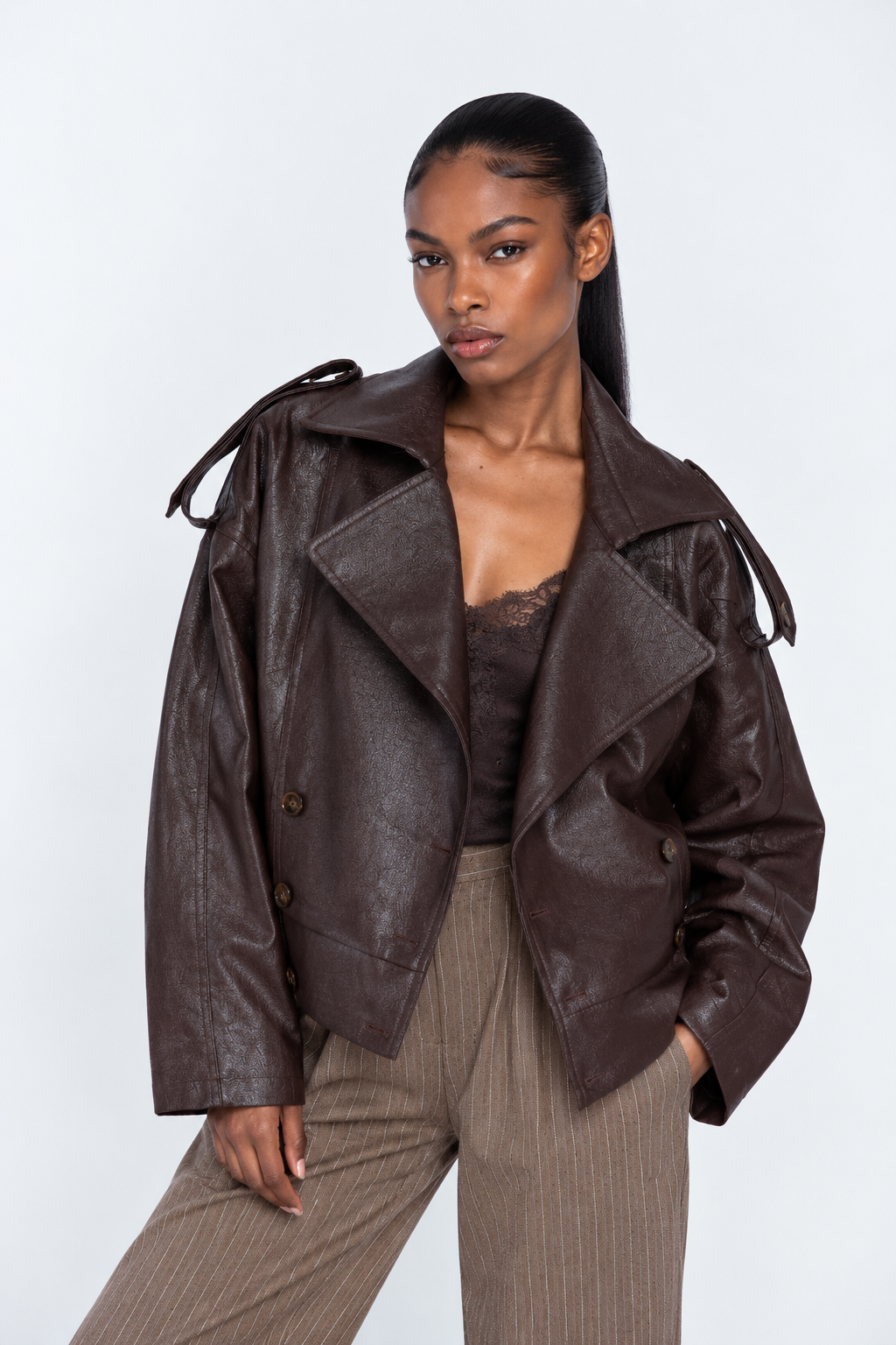 A woman modeling a dark brown faux-leather moto jacket with shoulder tie details, paired with beige pinstripe trousers, against a light gray studio background.