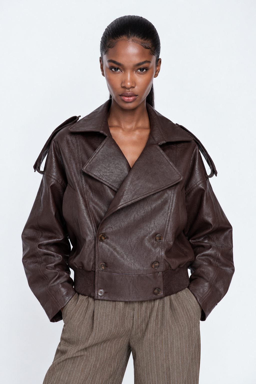 A model wearing a dark brown faux-leather double-breasted jacket with shoulder epaulets and brown pinstripe trousers, shot against a light gray studio background.