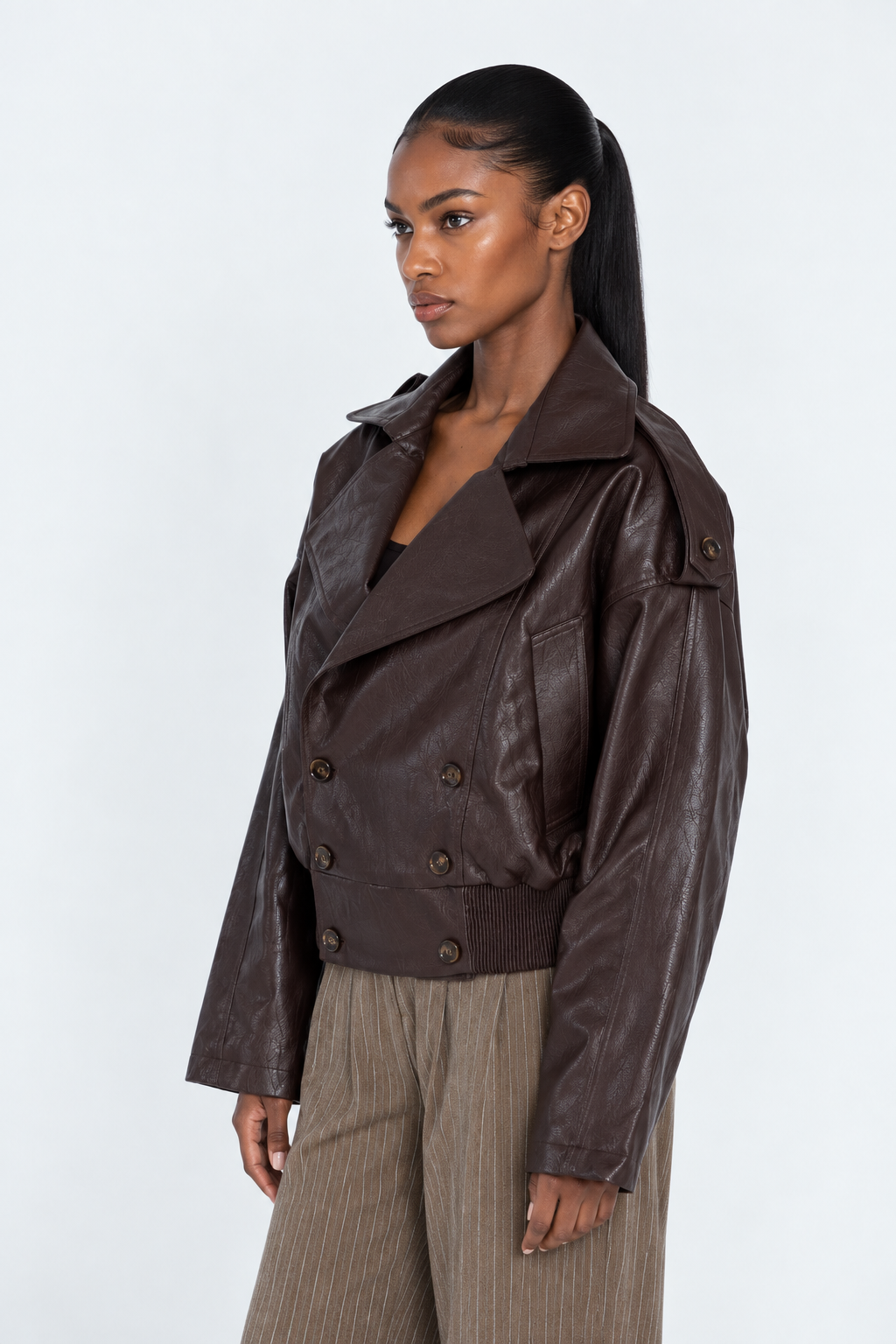 A woman modeling a dark brown faux-leather double-breasted jacket with a wide collar, paired with beige pinstripe trousers, against a plain white studio background.